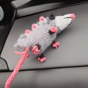 May include: A gray and pink crocheted opossum plush toy with a long tail. The toy is sitting on a black surface.