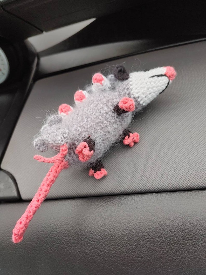 May include: A gray and pink crocheted opossum plush toy with a long tail. The toy is sitting on a black surface.