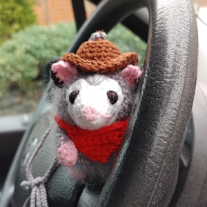 May include: A handmade crochet opossum wearing a brown cowboy hat and a red bandana. The opossum has gray fur, pink ears and paws, and black eyes. A gray string is attached to the opossum. The opossum is positioned in front of a steering wheel.