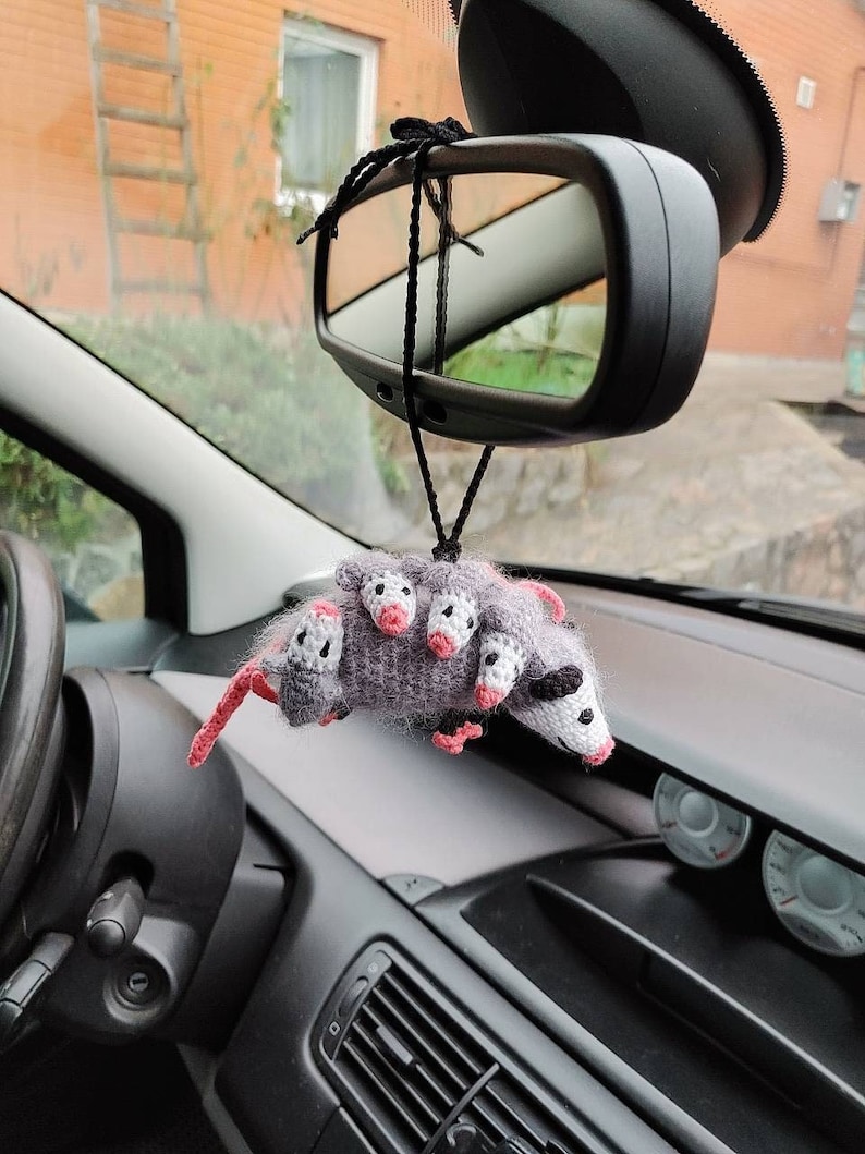 May include: A gray crocheted opossum with pink ears and nose hangs from the rearview mirror of a car. The opossum has several smaller opossums on its back.