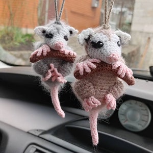 May include: Two handmade crochet possum car charms. One is gray, the other is brown, both with pink paws and tails. They are hanging from a string, with a brown stick in their arms.