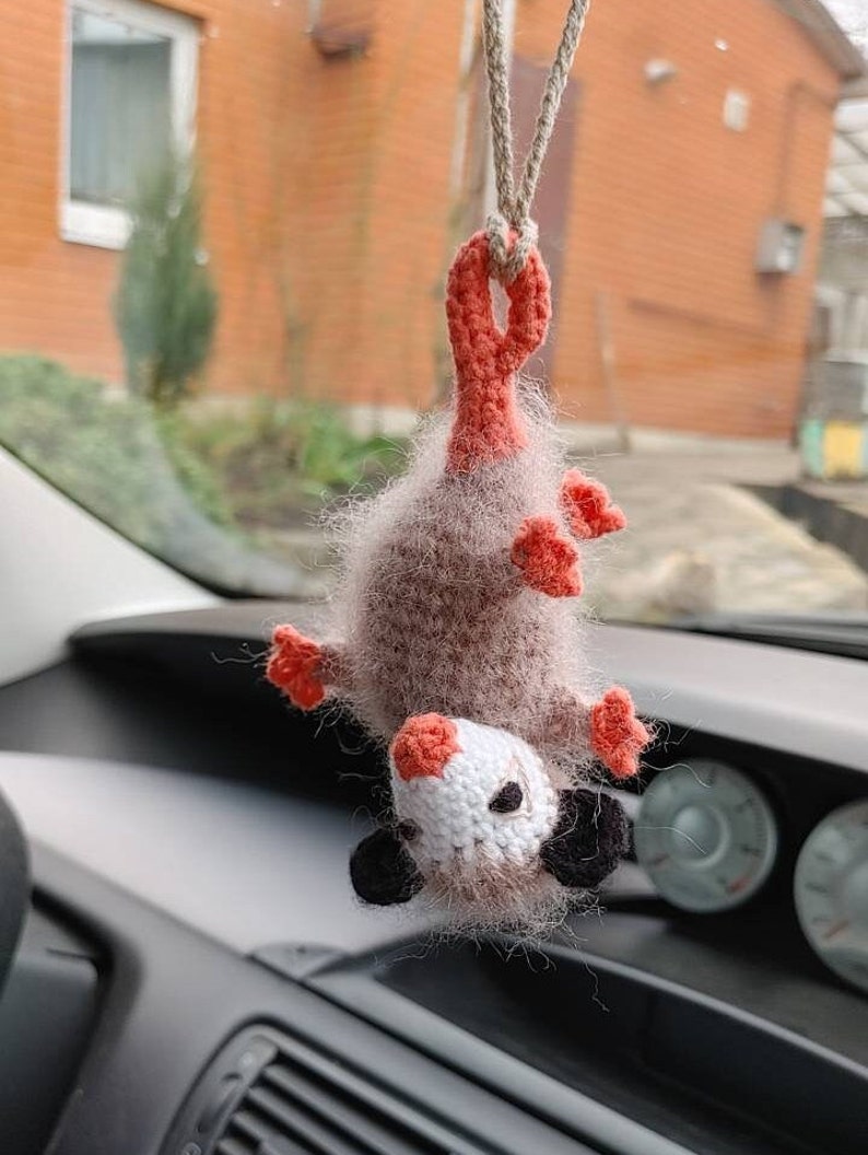 May include: A crocheted opossum hanging from a rear view mirror in a car. The opossum is brown and grey with pink ears and a white face. It has black eyes and a long tail.
