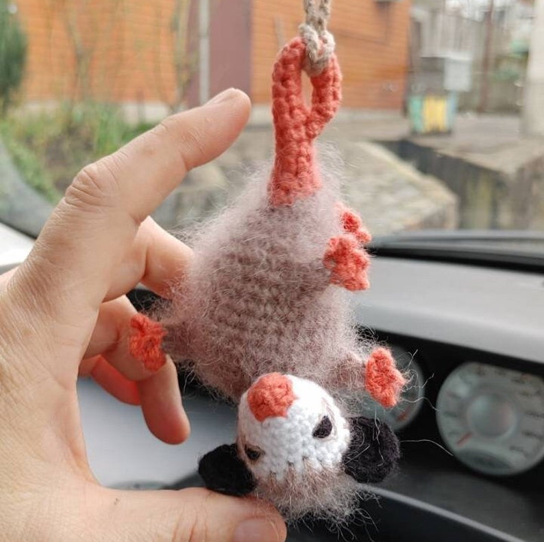 May include: A crocheted plush toy shaped like an opossum, hanging from a string. The opossum is brown and white with pink ears and feet. It has a long tail and a small, pointed nose.