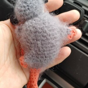 May include: A grey crocheted monkey toy with orange legs and tail. The toy is being held in a person's hand.