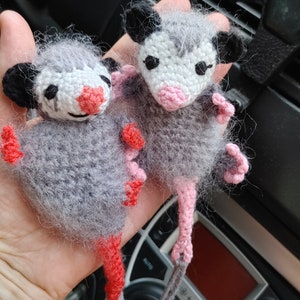 May include: Two crocheted opossum plushies with pink and red accents. The opossums are gray and white with black eyes and pink noses. They are hanging from a gray string.