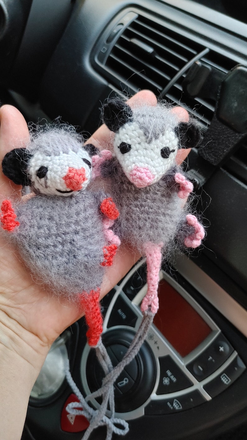 May include: Two crocheted opossum plushies with pink and red accents. The opossums are gray and white with black eyes and pink noses. They are hanging from a gray string.