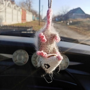 May include: A crocheted opossum hanging from a rearview mirror in a car. The opossum is grey and white with pink ears and paws.