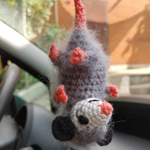 May include: A grey and white crocheted opossum hanging from a rear view mirror in a car. The opossum has pink ears and feet.