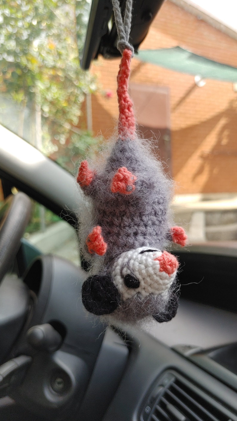 May include: A grey and white crocheted opossum hanging from a rear view mirror in a car. The opossum has pink ears and feet.
