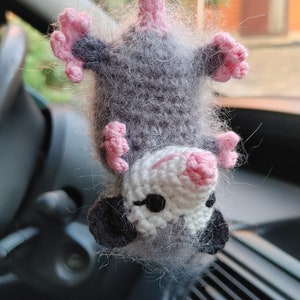 May include: A gray and white crocheted opossum hanging from a rearview mirror in a car. The opossum has pink ears and a pink nose.