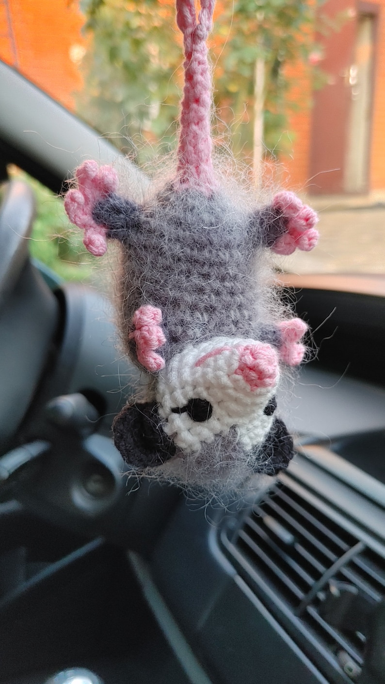 May include: A gray and white crocheted opossum hanging from a rearview mirror in a car. The opossum has pink ears and a pink nose.