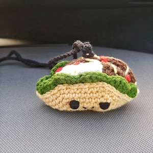Op de afbeelding: Een gehaakte taco sleutelhanger met een groene sla, bruin vlees en witte kaas vulling. De taco heeft een schattig gezicht met zwarte ogen.