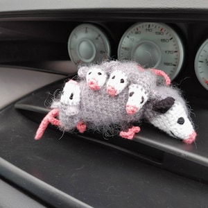 May include: A gray and white crocheted opossum toy with pink ears and nose. The toy is sitting on the dashboard of a car.