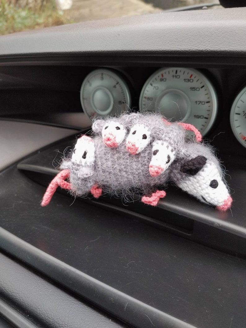 May include: A gray and white crocheted opossum toy with pink ears and nose. The toy is sitting on the dashboard of a car.