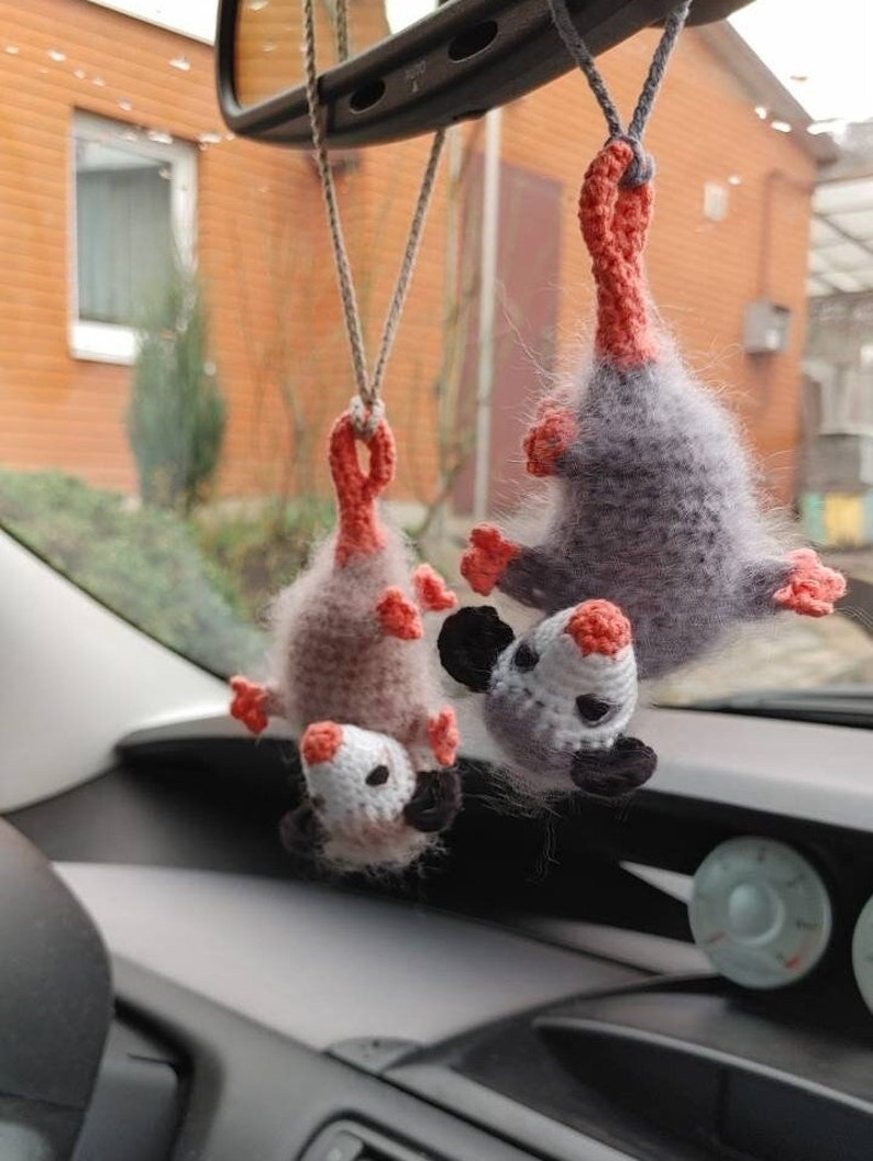 May include: Two crocheted opossum plushies hanging from a rear view mirror in a car. The opossums are grey and brown with pink ears and noses.