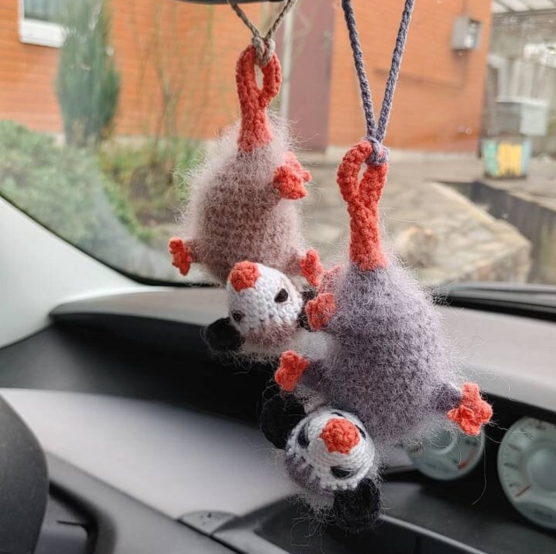 May include: Three crocheted opossum plushies hanging from a rear view mirror in a car. The opossums are grey and white with orange ears and noses.