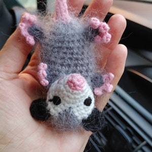 May include: A small gray and pink crocheted opossum with black eyes and a pink nose. The opossum is hanging upside down in a hand.