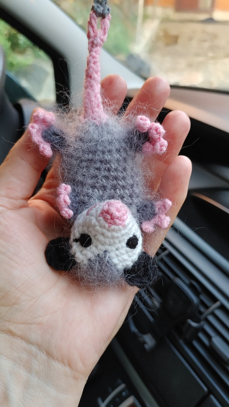 May include: A small gray and pink crocheted opossum with black eyes and a pink nose. The opossum is hanging upside down in a hand.