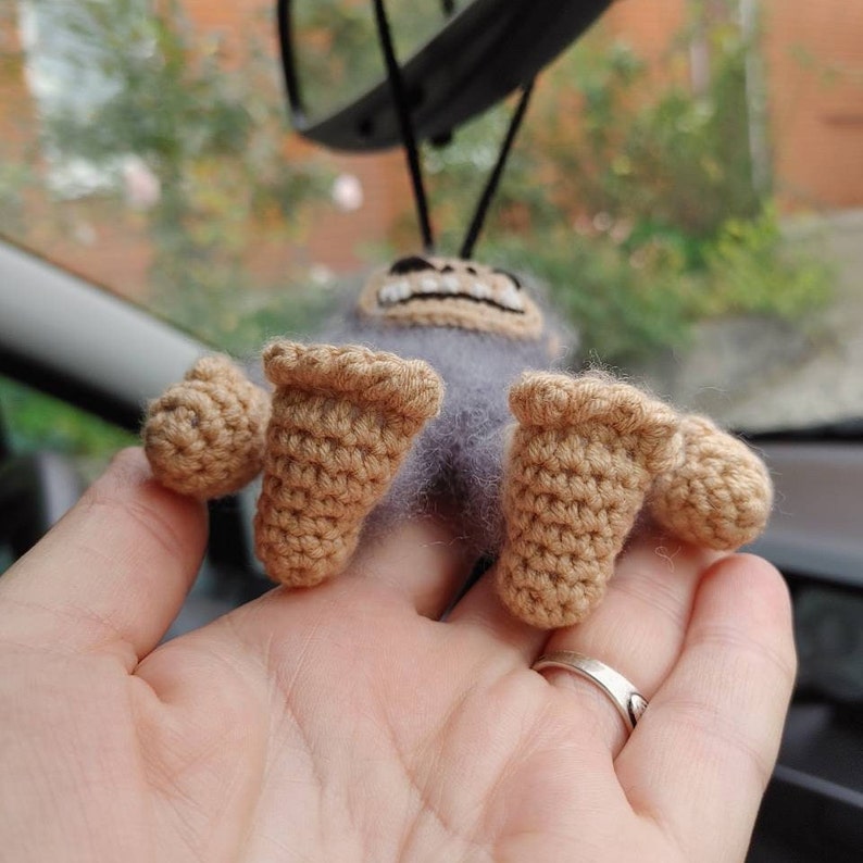 Puede incluir: Un juguete de peluche de crochet con forma de mono con una amplia sonrisa. El mono es gris con manos y pies de crochet marrones. El juguete est&aacute; colgado de un espejo retrovisor en un coche.