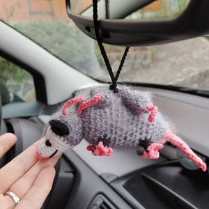 May include: A gray and pink crocheted opossum hanging from a rearview mirror in a car. The opossum has pink ears and feet.