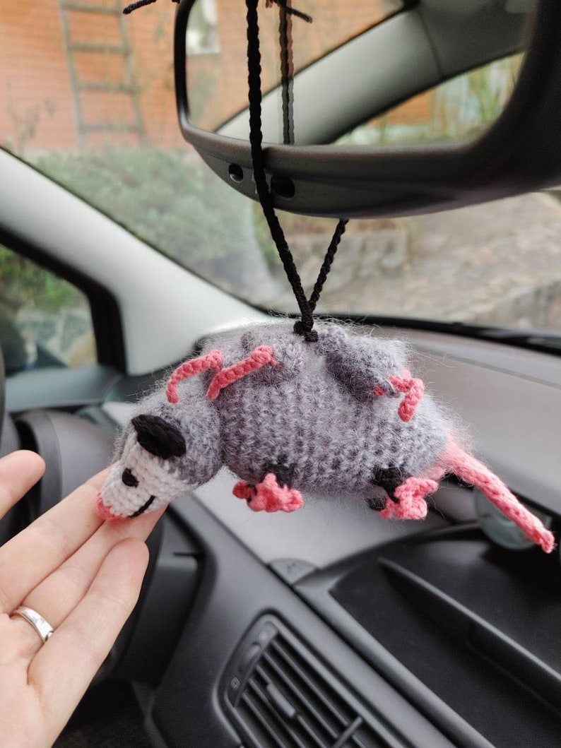 May include: A gray and pink crocheted opossum hanging from a rearview mirror in a car. The opossum has pink ears and feet.