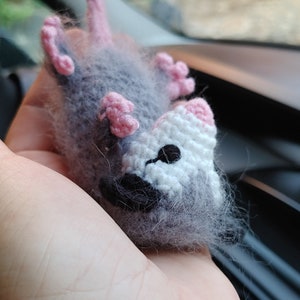 May include: A gray and white crocheted plush toy resembling a possum with pink ears and a black nose. The toy is hanging from a string.