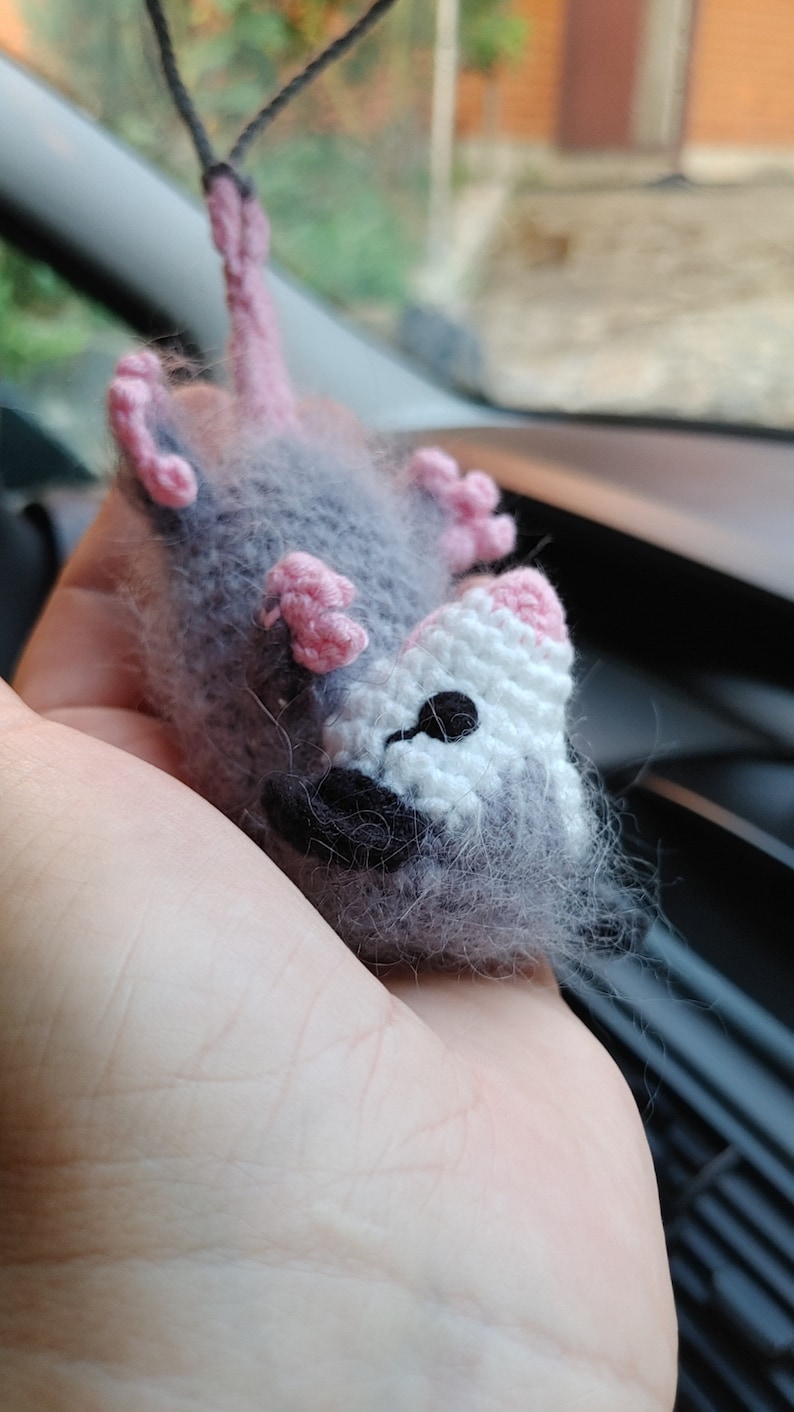 May include: A gray and white crocheted plush toy resembling a possum with pink ears and a black nose. The toy is hanging from a string.