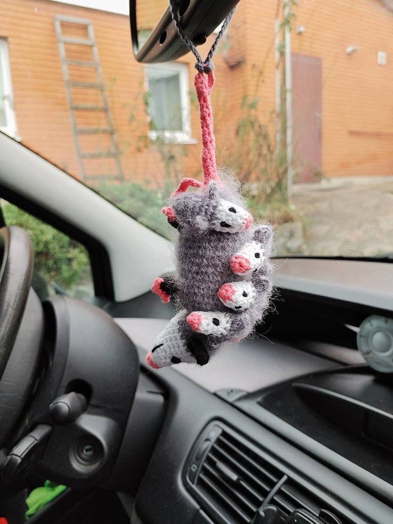 May include: A gray and white crocheted opossum hanging from a rearview mirror in a car. The opossum has pink accents and is holding onto a pink string.