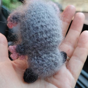 May include: A gray and pink knitted opossum plush toy with a long tail. The toy is held in a person's hand.