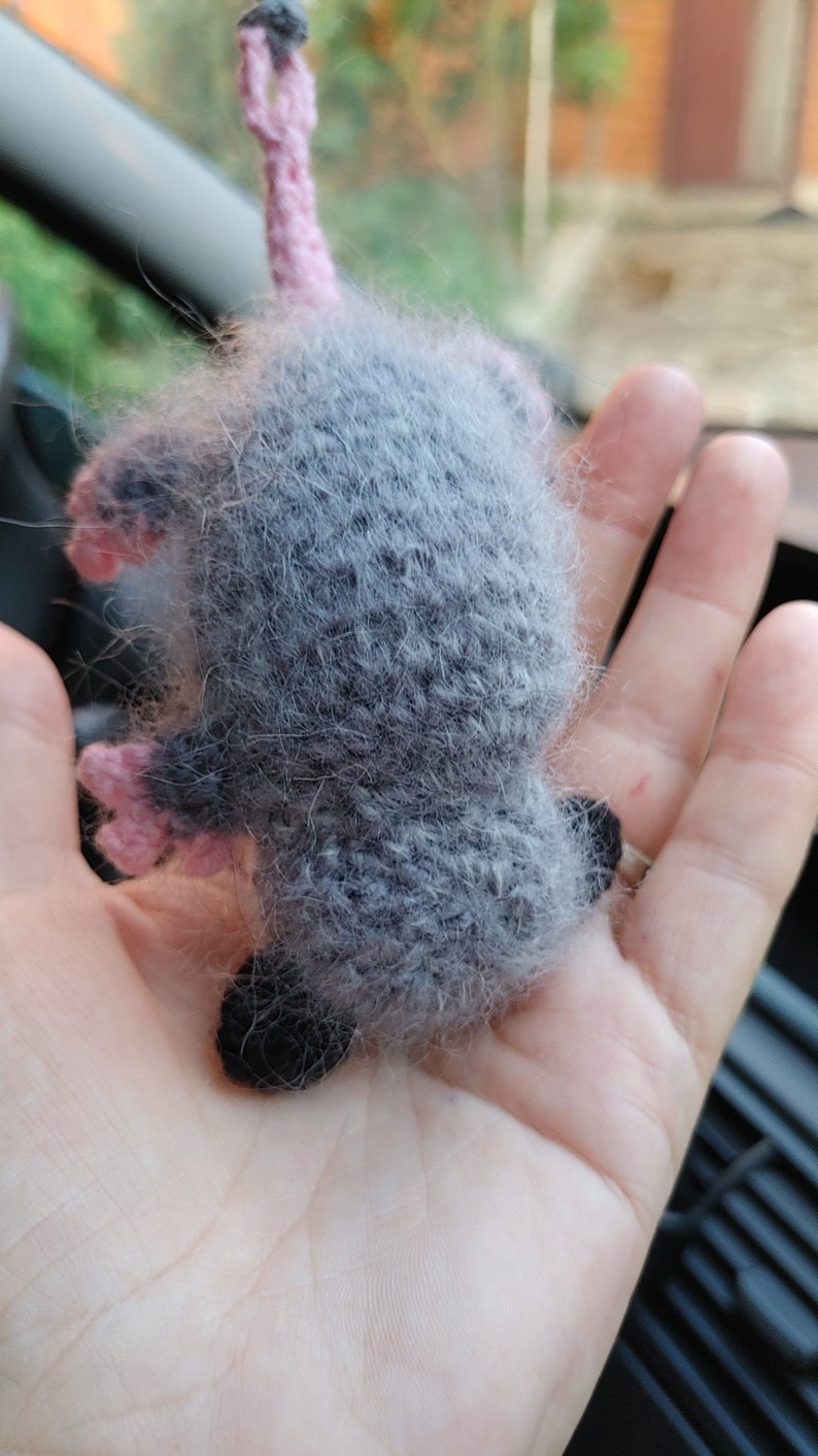 May include: A gray and pink knitted opossum plush toy with a long tail. The toy is held in a person's hand.