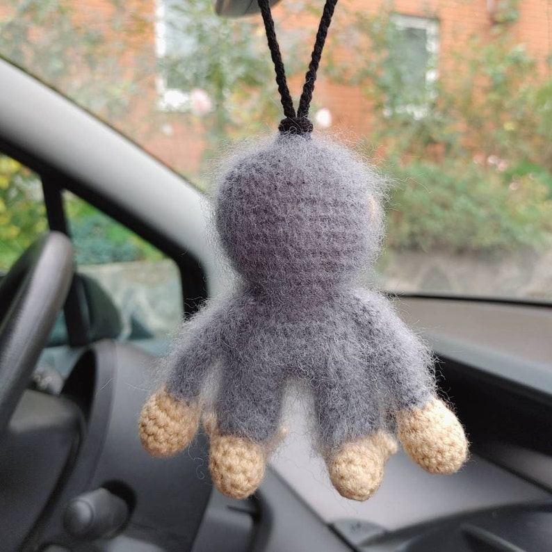 Puede incluir: Un mono de crochet gris colgando del espejo retrovisor de un coche. El mono tiene una textura peluda y pies beige.