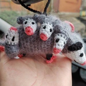 May include: A gray crocheted opossum with four heads, each with pink noses and white eyes. The opossum is hanging from a black string.