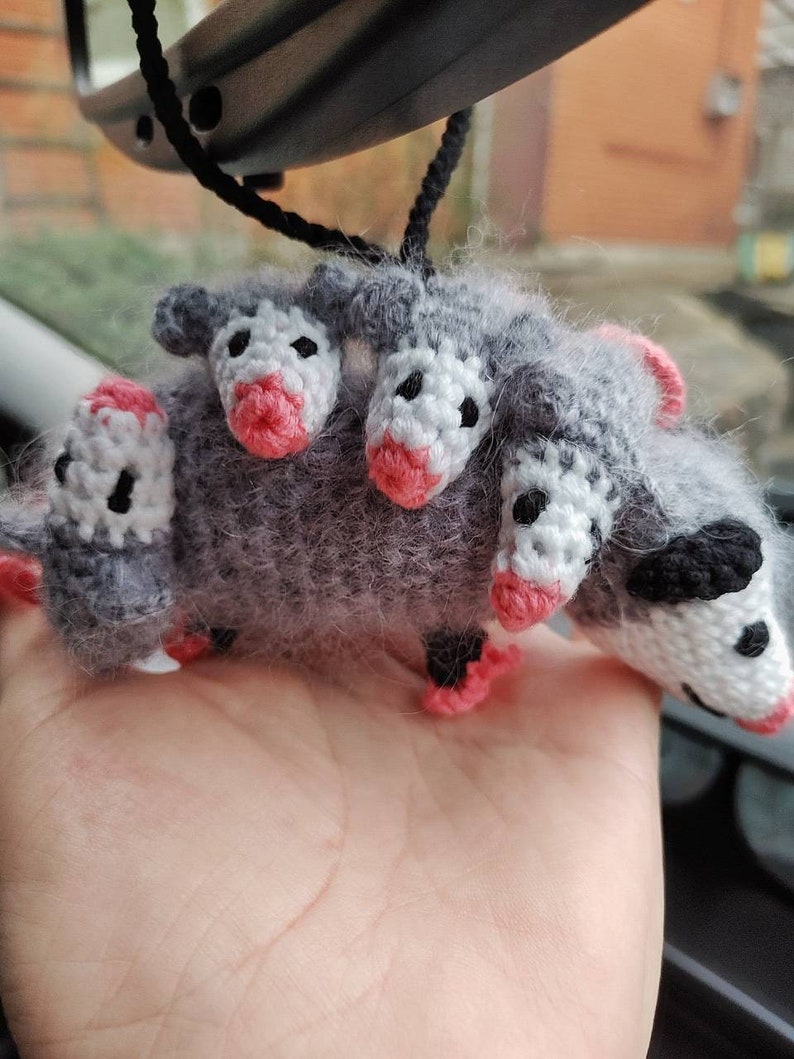May include: A gray crocheted opossum with four heads, each with pink noses and white eyes. The opossum is hanging from a black string.
