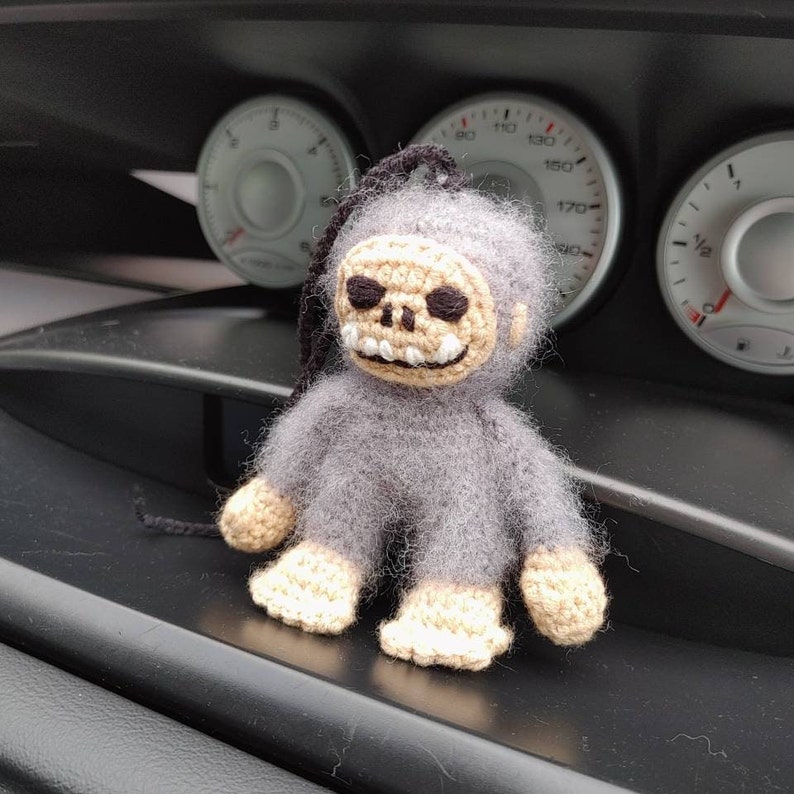 Puede incluir: Un mono de crochet gris con una cara de calavera y manos y pies de crochet blanco. El mono est&aacute; sentado sobre una superficie negra.