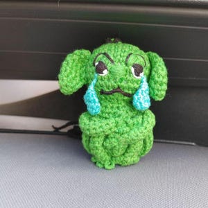 May include: A small, green, crocheted creature with floppy ears and a sad expression. It features two large, light blue, teardrop-shaped accents stitched under its black-embroidered eyes, with a small black mouth and crossed arms.