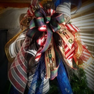 May include: A Christmas tree topper with a large bow made of red, white, and green plaid ribbon. The bow is surrounded by various types of mesh ribbon, including black and white striped, red and white striped, gold, and blue.