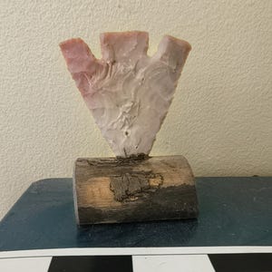 Peut inclure: Une pointe en pierre rose pâle à trois branches avec une texture écaillée, fixée solidement dans un morceau de bois cylindrique horizontal avec de l'écorce visible. Une échelle graduée noire et blanche est visible en bas du cadre.