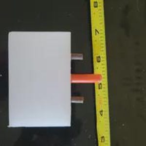 May include: A rectangular white block with three protruding prongs, including one orange center prong and two silver side prongs, positioned next to a yellow measuring tape showing a length of approximately 2.5 inches for the white block.