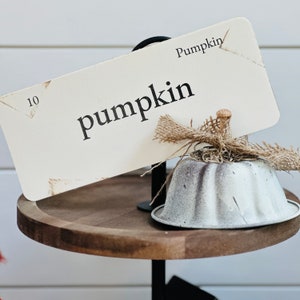 May include: A white wooden sign with the word "pumpkin" written in black lettering. The sign is attached to a black metal stand with a white metal pumpkin-shaped container. The sign also has the number "10" written on it.