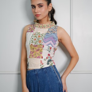 May include: A sleeveless white top with a patchwork design featuring floral and geometric patterns. The top has a high neckline and is paired with a blue denim skirt.