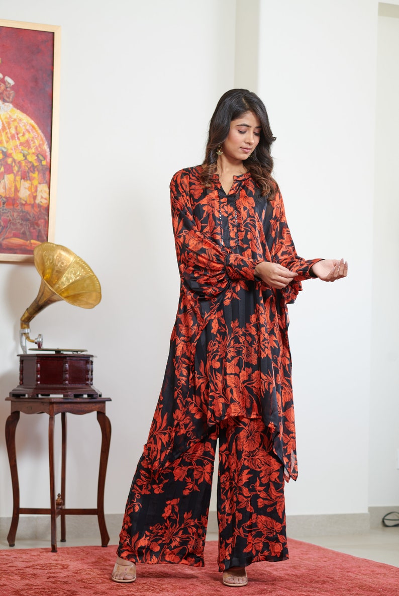 Pu&ograve; includere: Una donna indossa una blusa e pantaloni abbinati con stampa floreale nera e arancione. La blusa ha una silhouette lunga e fluida e una scollatura a V. I pantaloni sono a gamba larga e hanno una vestibilit&agrave; rilassata.