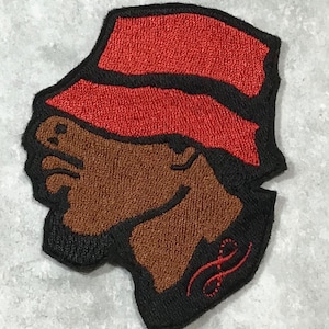May include: Embroidered patch of a person's head wearing a red bucket hat. The person's skin is brown and the patch has a black outline. The patch has a red swirl design in the bottom right corner.