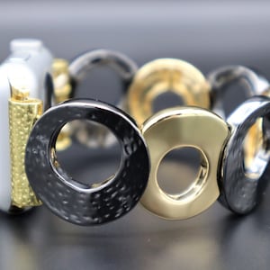 Puede incluir: Una pulsera de reloj de metal negro y dorado con un acabado texturizado. La pulsera está compuesta por círculos entrelazados.
