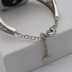 May include: A silver watch band with a decorative, filigree design. The band has a clasp closure and a small, round charm.