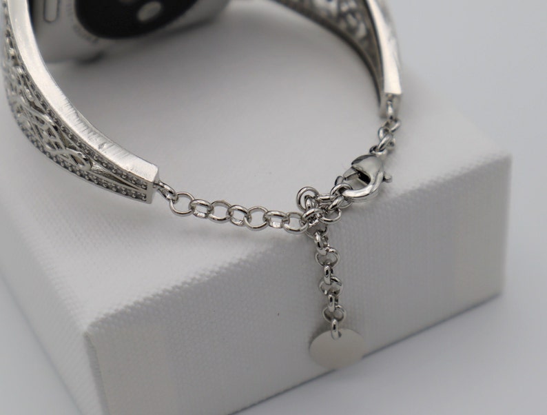 May include: A silver watch band with a decorative, filigree design. The band has a clasp closure and a small, round charm.