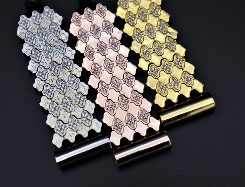 May include: Three metal watch bands with a geometric pattern and clear stones. The bands are silver, rose gold, and gold.