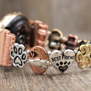 Puede incluir: Una pulsera de metal multicolor con abalorios de huellas de pata y un dije de coraz&oacute;n que dice "BEST FRIEND". La pulsera tiene una mezcla de elementos de color cobre, plata y oro, y est&aacute; sobre una superficie de madera.