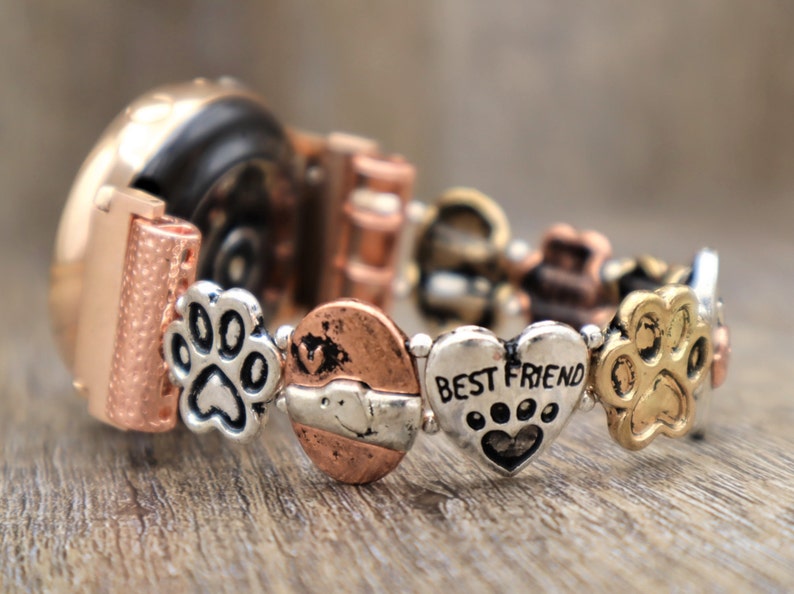 Puede incluir: Una pulsera de metal multicolor con abalorios de huellas de pata y un dije de coraz&oacute;n que dice "BEST FRIEND". La pulsera tiene una mezcla de elementos de color cobre, plata y oro, y est&aacute; sobre una superficie de madera.