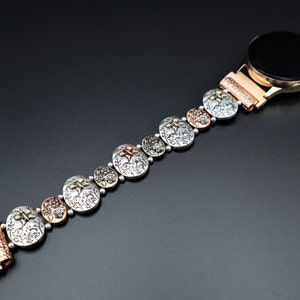 Pode incluir: Uma pulseira de rel&oacute;gio de prata, cobre e ouro com um design floral. A pulseira &eacute; composta por elos redondos individuais com um padr&atilde;o floral em relevo. A pulseira est&aacute; presa a um smartwatch com um mostrador preto.