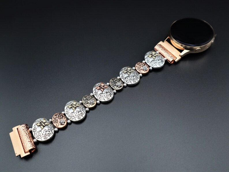 Pode incluir: Uma pulseira de rel&oacute;gio de prata, cobre e ouro com um design floral. A pulseira &eacute; composta por elos redondos individuais com um padr&atilde;o floral em relevo. A pulseira est&aacute; presa a um smartwatch com um mostrador preto.